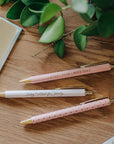 Three pens with motivational quotes on a wooden surface with green leaves.
