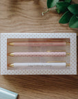 Set of three pens in a polka dot box on a wooden surface with green leaves.