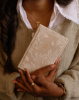 Person holding a beige Bible with floral designs, wearing a gold cross necklace and bracelet.