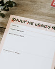 Notepad with 'Daily He Leads Me' text on a wooden surface
