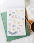 Green notebook with floral stickers on a white surface next to an open book and a cup of coffee.