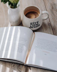 Open book on a wooden surface with a 'give me Jesus' mug and a vase with flowers.