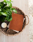 Wicker basket with a brown leather notebook, green plant, and candle on a wooden surface.