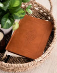 Brown leather notebook with embossed design in a woven basket with a candle and plant.