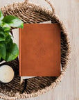 Brown leather notebook with tree design in a woven basket with a plant and candle on a wooden surface