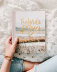 Person holding a book titled 'The Lord's Prayer' with a scenic cover against a textured white background.