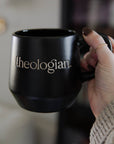 Black mug with 'theologian' text held by a person in a blurred indoor setting