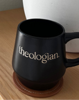 Black mug with 'theologian' on a wooden surface next to a white lamp.
