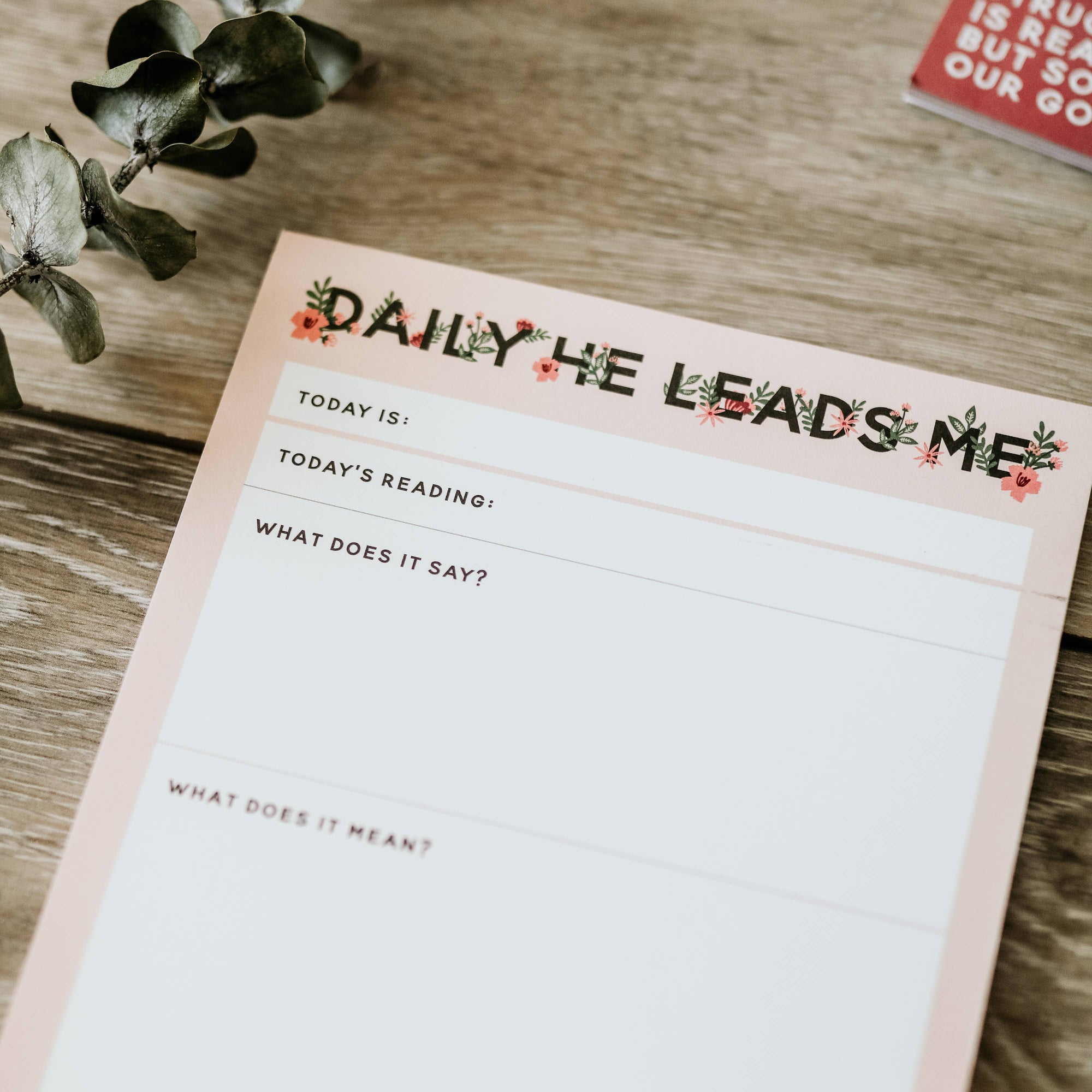 Notepad with 'Daily He Leads Me' text on a wooden surface
