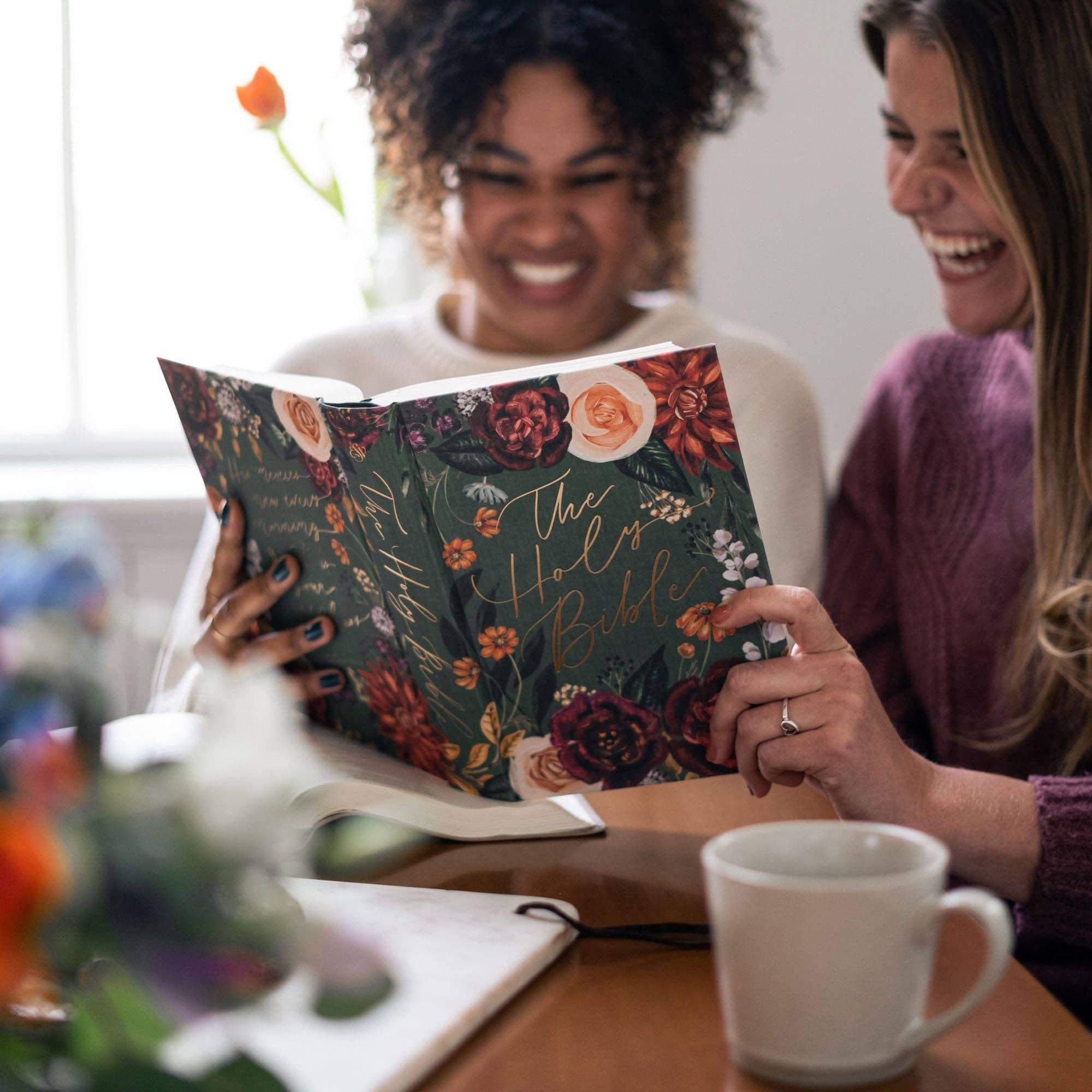 Two women reading a floral-covered Bible together, with a cup on a table in the foreground.