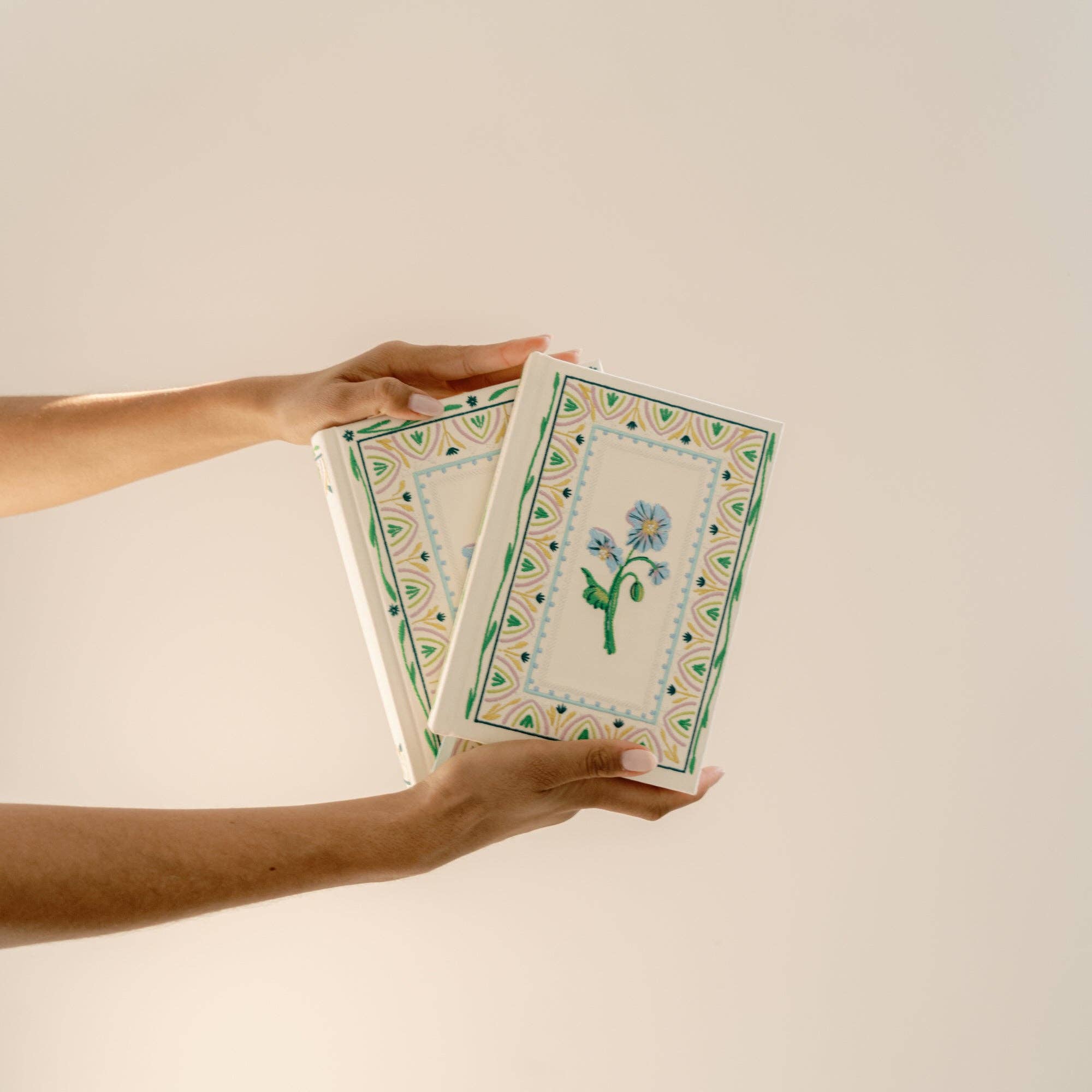 Two hands holding two notebooks with floral designs on a beige background