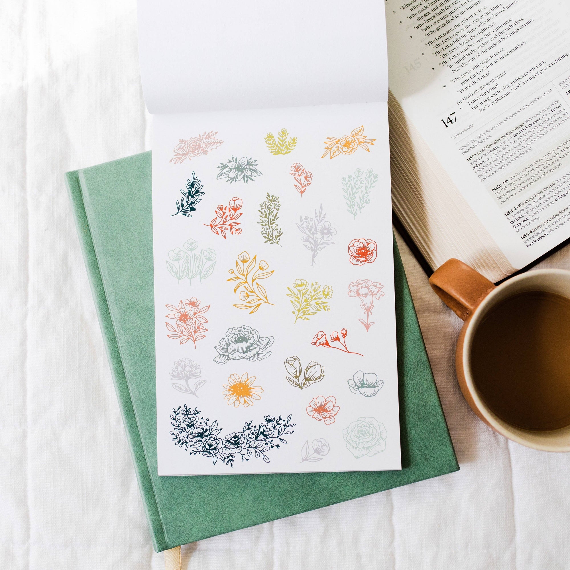 Green notebook with floral stickers on a white surface next to an open book and a cup of coffee.