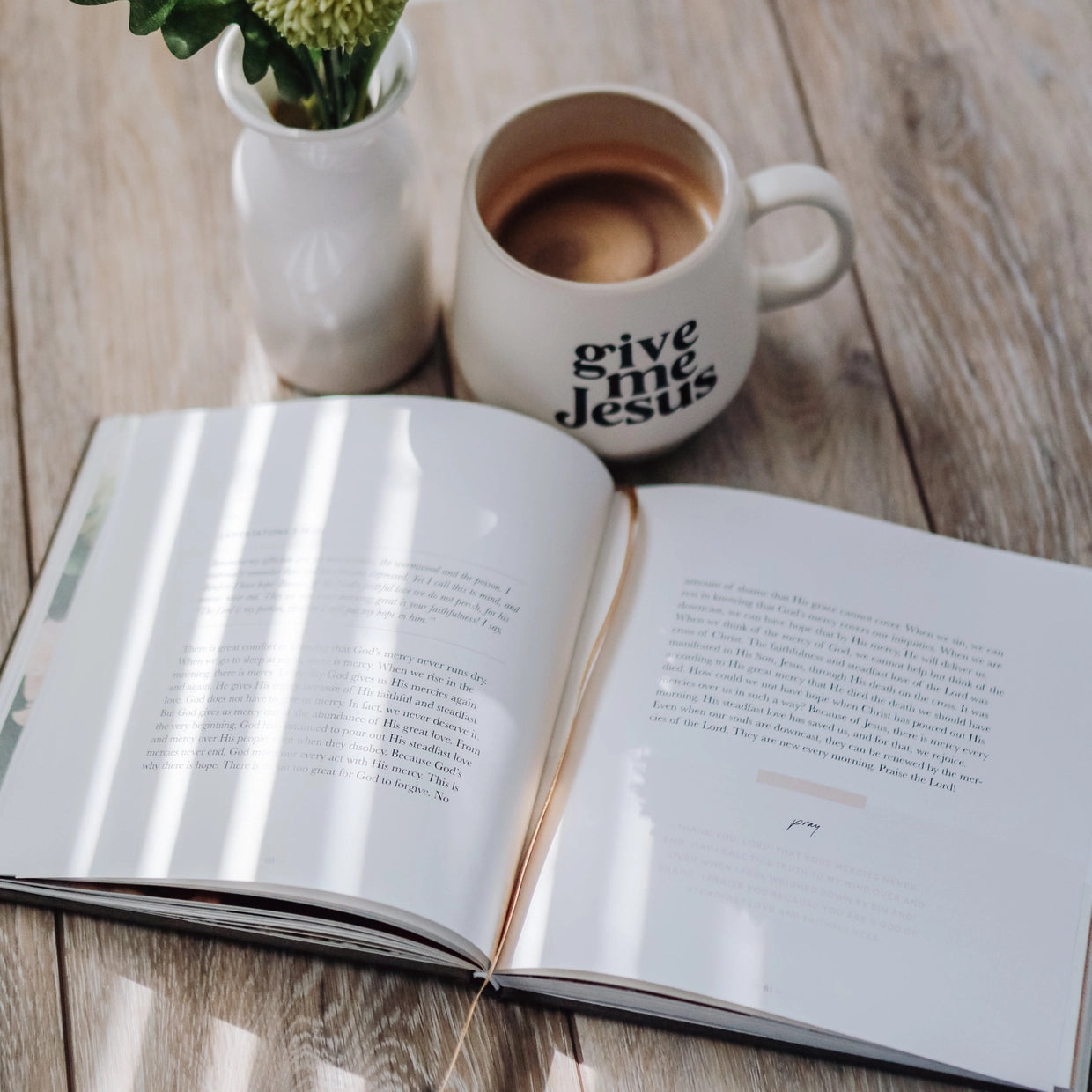 Open book on a wooden surface with a 'give me Jesus' mug and a vase with flowers.