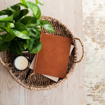 Wicker basket with a brown leather notebook, green plant, and candle on a wooden surface.