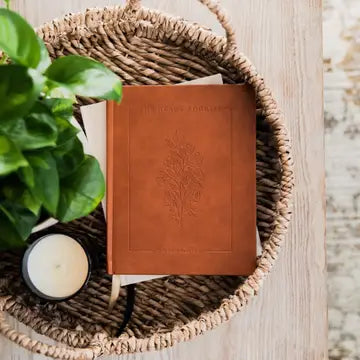 Brown leather notebook with tree design in a woven basket with a plant and candle on a wooden surface