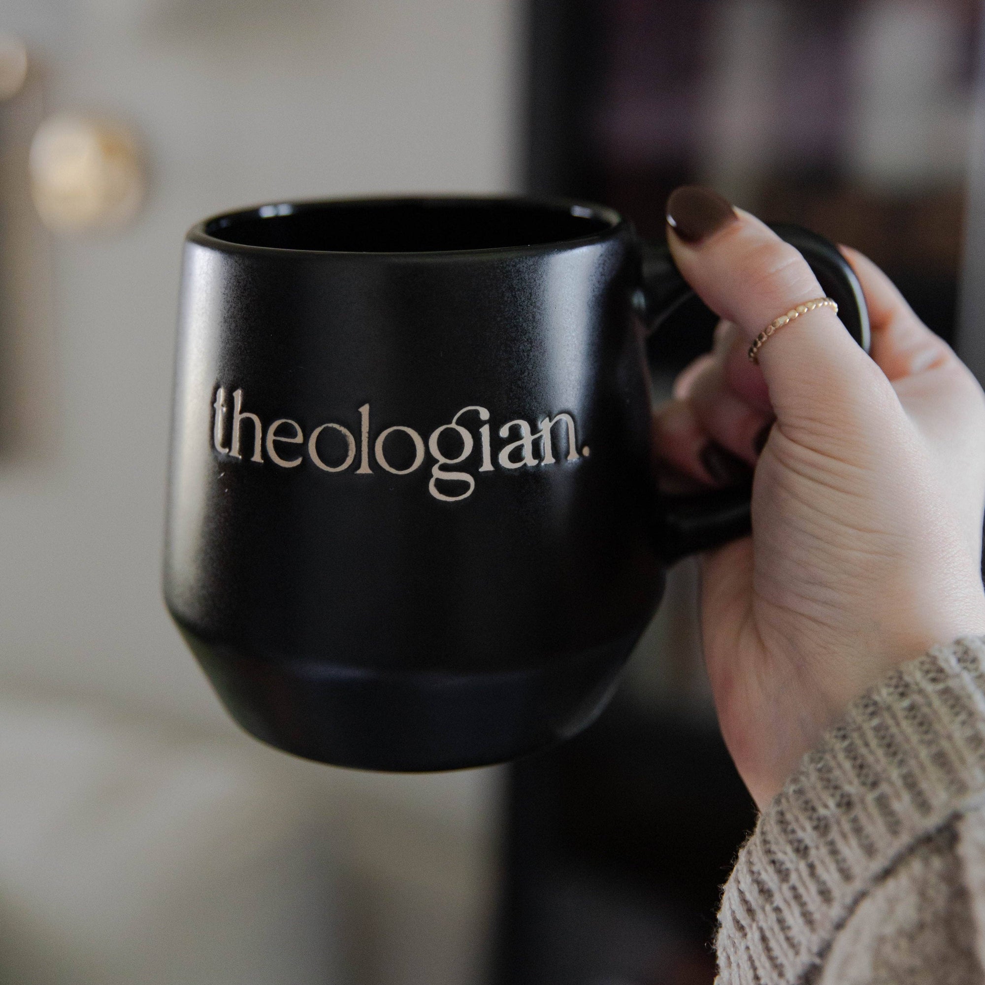 Black mug with 'theologian' text held by a person in a blurred indoor setting