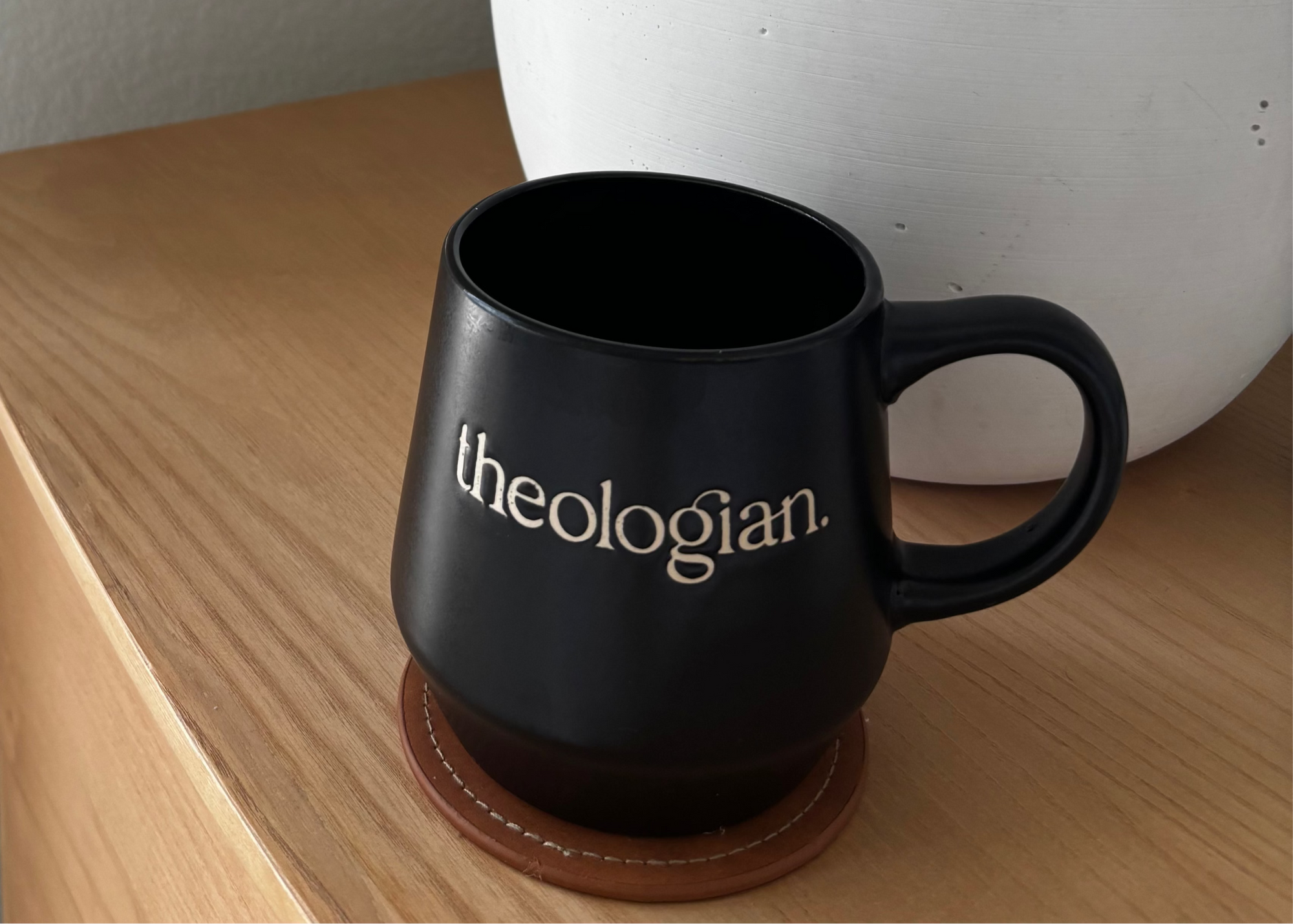 Black mug with 'theologian' on a wooden surface next to a white lamp.